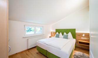 A cozy bedroom with a green headboard and white bedding. Large windows let in plenty of light, and the wooden floors add a warm touch.