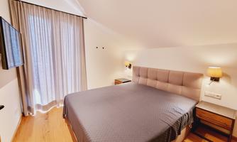 A modern bedroom with a large bed and a bright atmosphere. The windows are adorned with curtains and a TV is mounted on the wall.