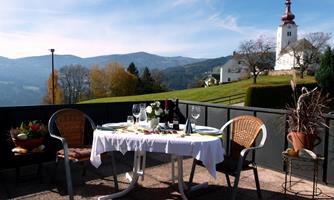 A beautiful table with table decorations and glasses is set on a terrace overlooking the mountains. In the background, a church and a picturesque landscape can be seen.