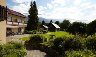A picturesque landscape with a well-kept garden, surrounded by trees and mountains. In the background, buildings and a clear blue sky are visible.