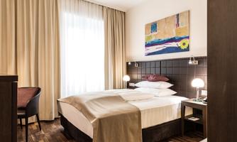 A modern hotel room with a double bed, a cozy seating area, and a colorful piece of art on the wall. Generous windows let in plenty of light and create a bright atmosphere.