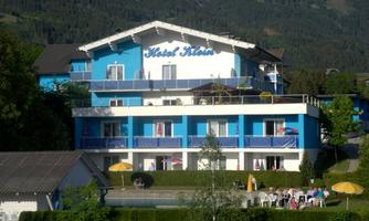 A blue hotel with several floors and balconies. In the foreground, there is a pool and some guests relaxing outdoors.
