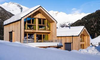Two modern wooden houses in a snowy mountain landscape. The mountains in the background are snow-covered and the sky is clear.
