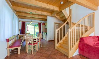 A bright living room with a seating area and a wooden dining table. In the background, there is a wooden staircase leading to the upper area.
