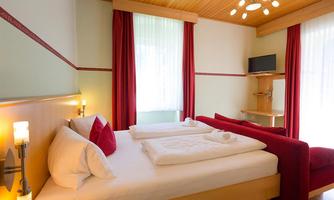 A cozy hotel room with a double bed and a red sofa. Bright curtains and modern furniture create an inviting atmosphere.