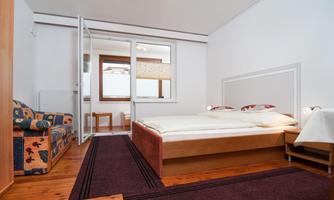 A cozy room with a double bed, a comfortable armchair, and a window that lets in daylight. The floor is wooden, and the room is simply but stylishly decorated.