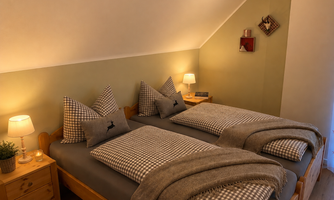 The image shows a cozy bedroom with two beds and checkered pillows. Gentle light and a warm color scheme create an inviting atmosphere.