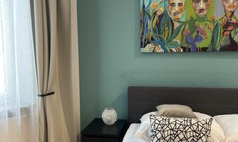 A cozy bedroom with a modern bed, a gray blanket, and a decorative cushion. A colorful artwork featuring faces and plant motifs hangs on the wall.