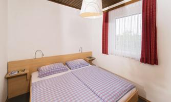 A cozy bedroom with a double bed and checkered bedspreads. The walls are light, and there are red curtains as well as a lamp above the bed.