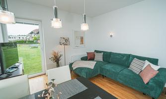 A modern living room with a green sofa and a dining table.  
Large windows let in plenty of light and provide a view of the garden.