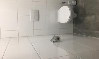 A modern bathroom with white tiles and a clean toilet. There is a toilet paper holder and a toilet cleaner.
