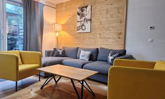 A modern living room with a blue couch and two yellow armchairs. The wooden table and wood paneling create a cozy atmosphere.
