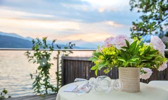 A stylishly set table with a floral arrangement and glasses overlooks a beautiful lake at sunset. In the background, mountains and a calm water surface can be seen.
