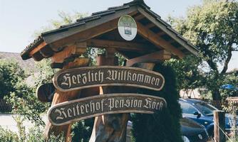 A rustic welcome sign with the inscription "Warm Welcome" and "Rider Farm Pension Böck". The sign is made of wood and surrounded by green nature.
