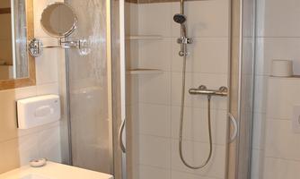 A modern shower cabin with clear glass walls in a practical bathroom. The walls are brightly tiled and there is a sink with a mirror.