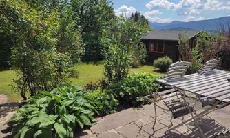 A beautiful garden with green plants and a clear blue sky. In the foreground, there is a table with chairs on a terrace.