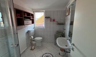 A modern bathroom with a shower, a toilet, and a sink. Large tiles and a window provide plenty of light.