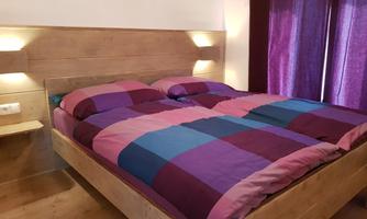 A modern bedroom with a double bed in a colorful checkered pattern. The walls are light, and the windows have dark curtains.