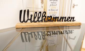 A welcome sign made of black metal and wood on a glass table. In the background, another sign with a saying is visible.