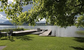 A quiet lake promenade with a wooden walkway and green grass. Thick trees provide shade and gentle hills are visible in the background.