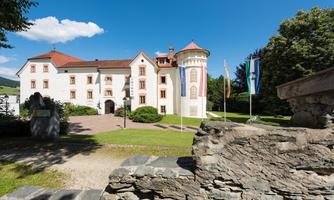 An elegant castle with a red roof and white walls in a green environment. The sun is shining and the sky is blue.