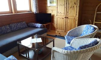 A cozy living room with wooden paneling. There is a couch, two rattan chairs, and a glass table, as well as a TV and windows with plenty of light.