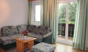 A cozy living room with a comfortable sofa and a wooden table. Through the windows, a green view of the balcony is visible.