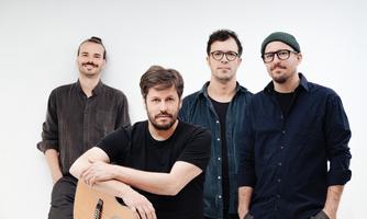 Eine Gruppe von vier Männern steht vor einem hellen Hintergrund. Einer der Männer hält eine Gitarre in den Händen.