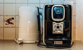 A modern espresso machine stands next to a kettle on a countertop. The kitchen has a bright, friendly atmosphere.
