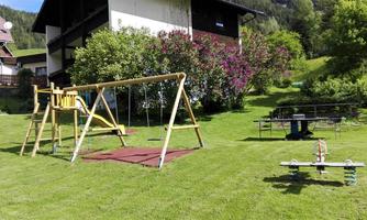 A playground with a swing, a seesaw, and a sandy ground in a green garden. In the background, there is a two-story house.