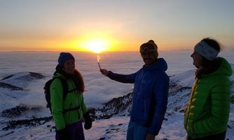 Three people are standing on a snow-covered mountain, enjoying the sunset. The view shows clouds beneath them and a clear sky.