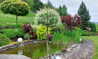 A beautiful garden with a pond, surrounded by colorful plants and decorative figures. In the background, there are some trees and a green lawn.