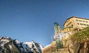 An impressive mountain landscape under a clear night sky. In the foreground stands a building, surrounded by majestic mountains and rocks.