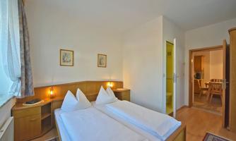 A simple bedroom with two single beds and wooden furniture. On the right side of the image is access to another room.