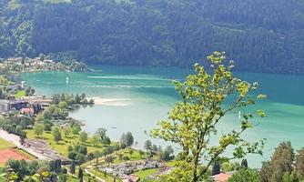 A beautiful lake surrounded by green trees and mountains. In the background, small houses and meadows can be seen.