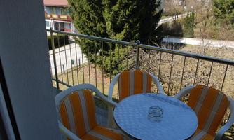 A balcony with two orange-striped chairs and a round table. In the background, trees and neighboring houses can be seen.