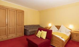 A cozy bedroom with a wooden wardrobe, a red carpet, and a double bed. The walls are painted in a warm shade of yellow, and there is a sofa with cushions.