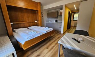 A cozy room with a folding bed and two single beds. The modern decor includes a table, chairs, and a kitchenette.