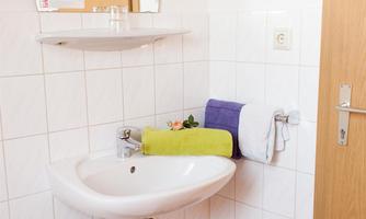A small, bright bathroom area with a white sink and a mirror. Towels in various colors are on a shelf, and there is a trash can in the corner.