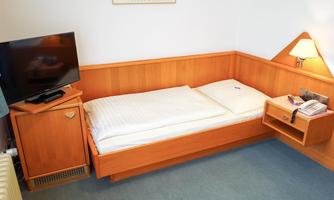 A simple single room with a bed, a television, and a nightstand. The walls are light and the furniture is made of wood.