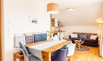 A modern dining kitchen with a large wooden table and comfortable chairs. In the background, there is a cozy seating area with a sofa and a small kitchen area.