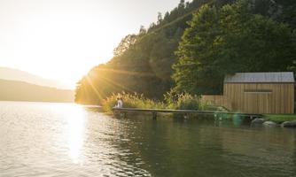A tranquil lake landscape at sunset. In the foreground, there is a pier and a modern wooden building, surrounded by trees.
