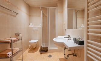 A modern bathroom with a shower, sink, and toilet. The walls are light-tiled, and there are towels on a shelf.