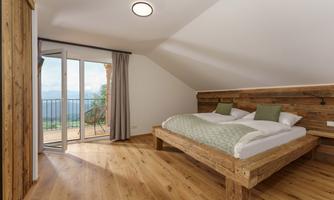 A modern bedroom with a large wooden bed and an open balcony. The bright walls and wooden floor create a cozy atmosphere.