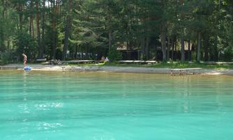 A clear, turquoise lake with a sandy shore. In the background, trees and loungers are visible.