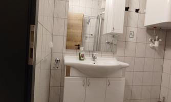 A modern bathroom with a white sink and a large mirror. The walls are tiled, and there is a shower in the background.