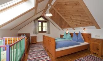 A cozy attic room with a wooden ceiling and two beds. In the room stands a children's bed and there is a colorful carpet.
