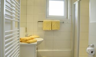A modern bathroom with white tiles and a window. Yellow towels are placed on the sink, and next to the shower area, there is a towel rack.