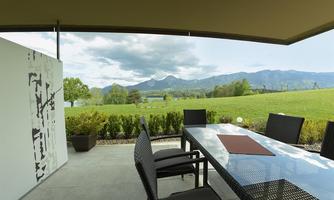 A modern terrace with a table and chairs, surrounded by a green meadow and mountains in the background. The sky is partly cloudy, creating a peaceful atmosphere.