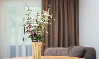 A pretty bouquet of flowers in a decorative vase is sitting on a wooden table. In the background, there are opaque curtains and a cozy couch visible.
